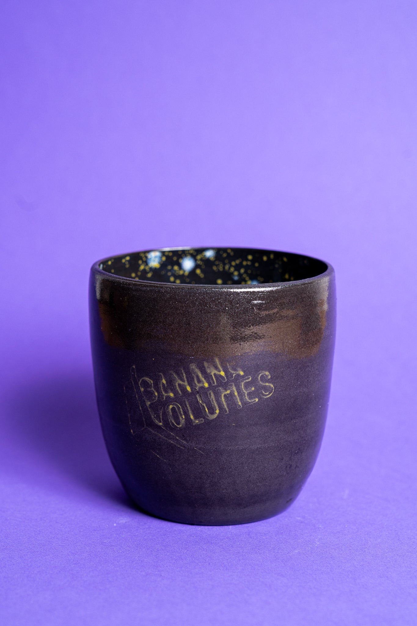 Eine schwarz-violette Tasse ohne Griff mit Banana Volumes Logo auf violettem Hintergrund.