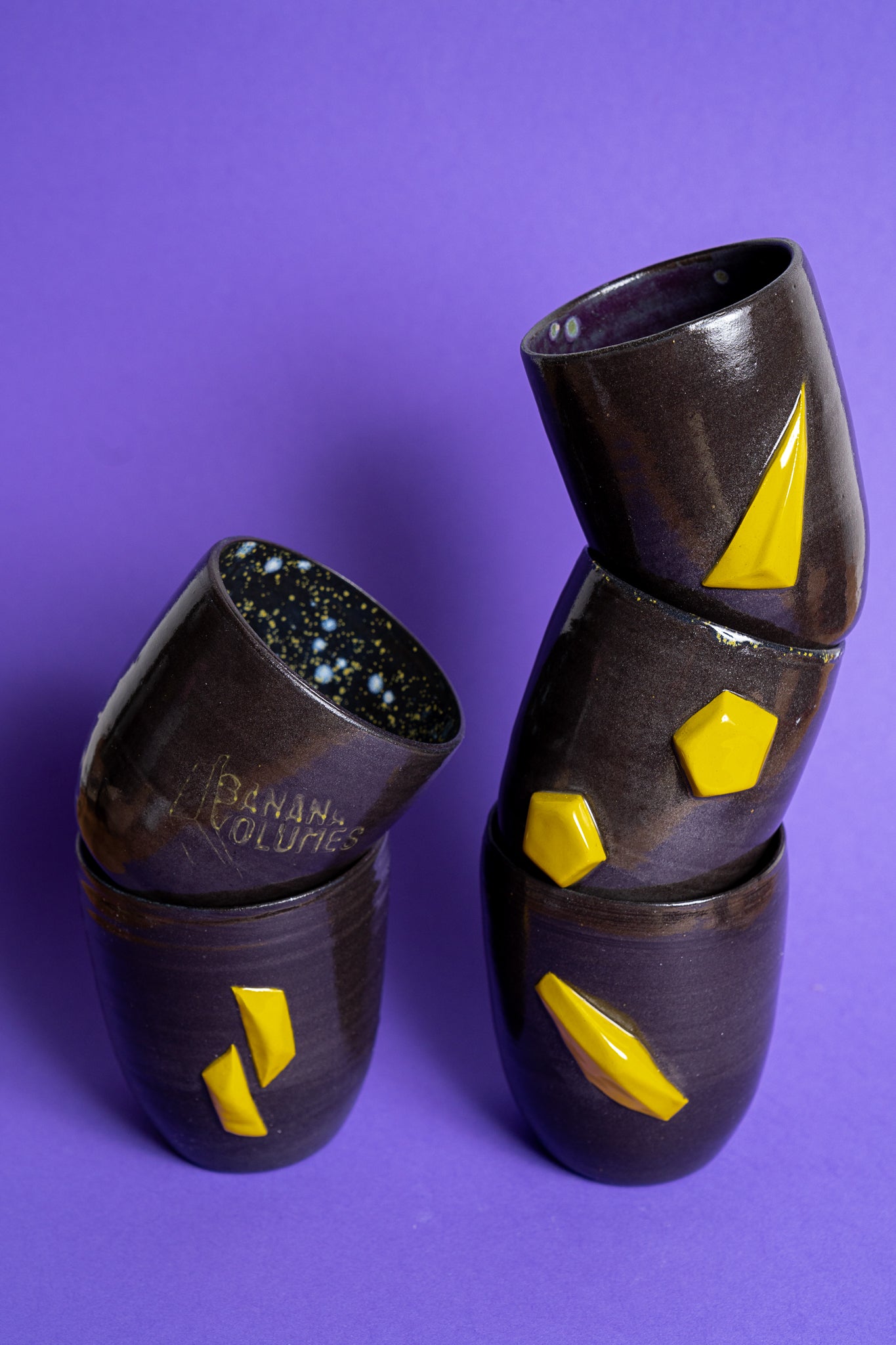 Fünf gestapelte, schwarz-violette Tassen ohne Griff mit Banana Volumes Logo und gelben Banana Volumes shapes auf violettem Hintergrund.