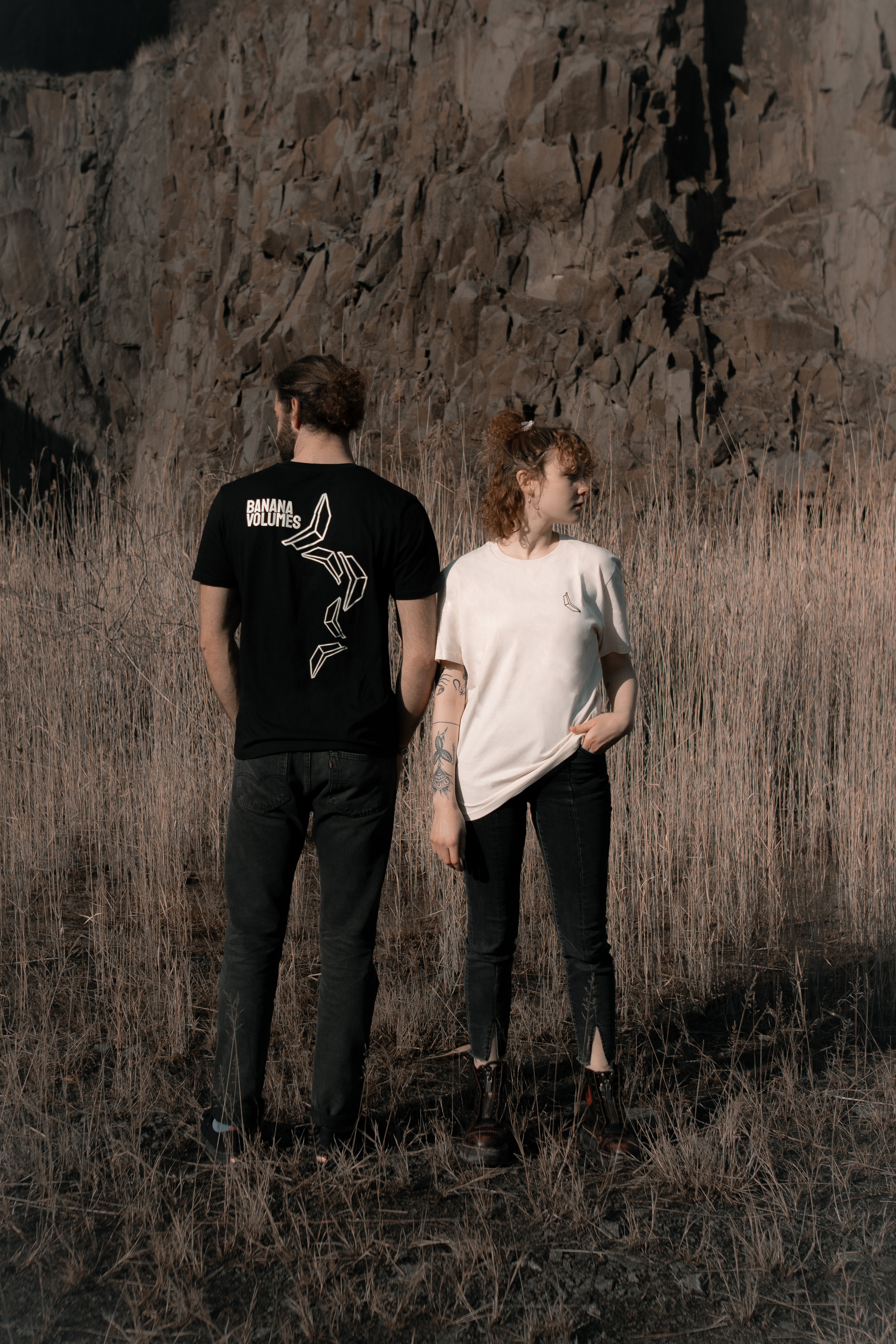 Eine Frau in einem weißen Banana Volumes T-Shirt und ein Mann in einem schwarzen Banana Volumes T-Shirt stehen nebeneinander in einem Feld vor einer Felswand.