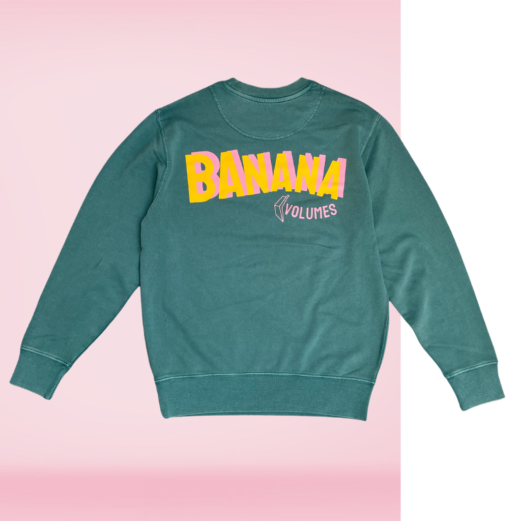 Die Rückseite eines grünen Sweaters mit Banana Volumes Print in gelb und rosa vor einem rosa-weißen Hintergrund.