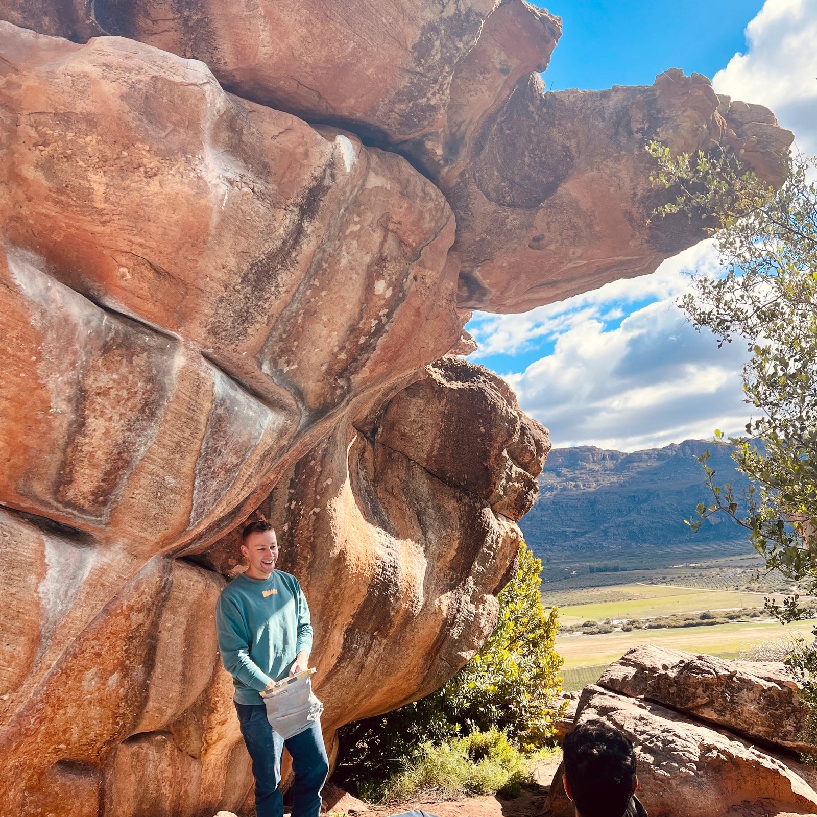 Ein Mann in einem grünen limited Edition Banana Volumes Sweater steht lachend vor einem Boulder in Rocklands, SA.
