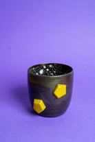 Eine schwarz-violette Tasse mit zwei gelben Banana Volumes Stacks shapes auf der Seite auf violettem Hintergrund.