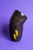 Zwei gestapelte schwarz-violette Tasse, eine mit gelbem Banana Volumes Logo und die andere mit zwei gelben Banana Volumes Boats shapes auf der Seite auf violettem Hintergrund.