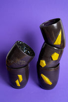 Fünf gestapelte, schwarz-violette Tassen ohne Griff mit Banana Volumes Logo und gelben Banana Volumes shapes auf violettem Hintergrund. 