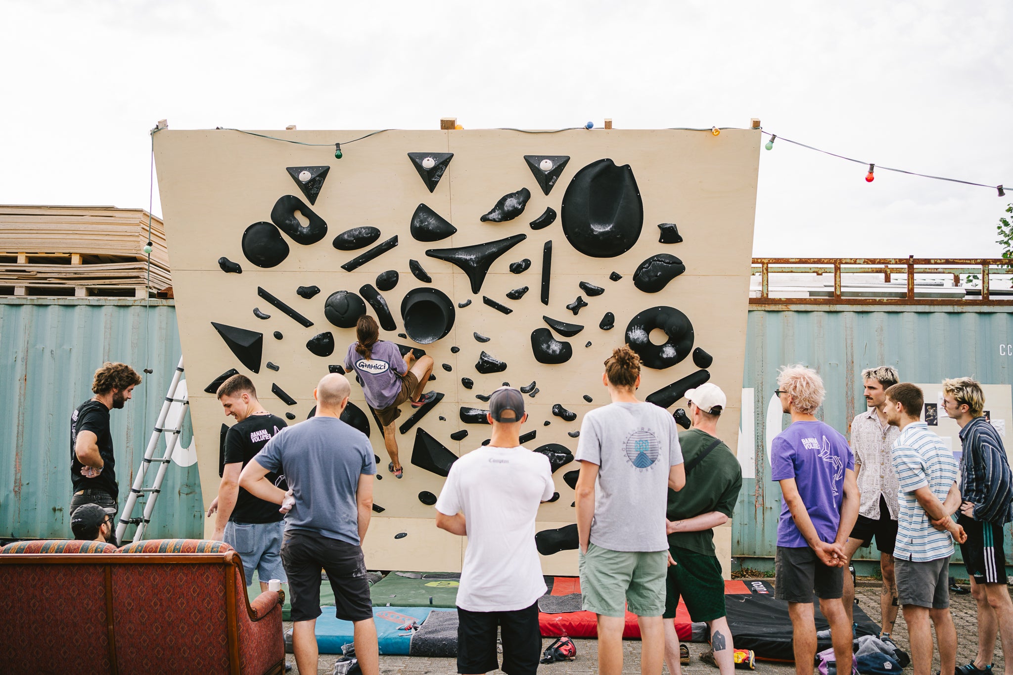 Zehn Leute stehen im Freien vor einer Boulderwand und schauen einer Person beim klettern zu. Regrab by Banana Volumes Community und Routenbau