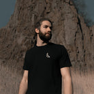 Ein Mann in einem schwarzen Banana Volumes T-Shirt steht in einem Feld vor einer Felswand.