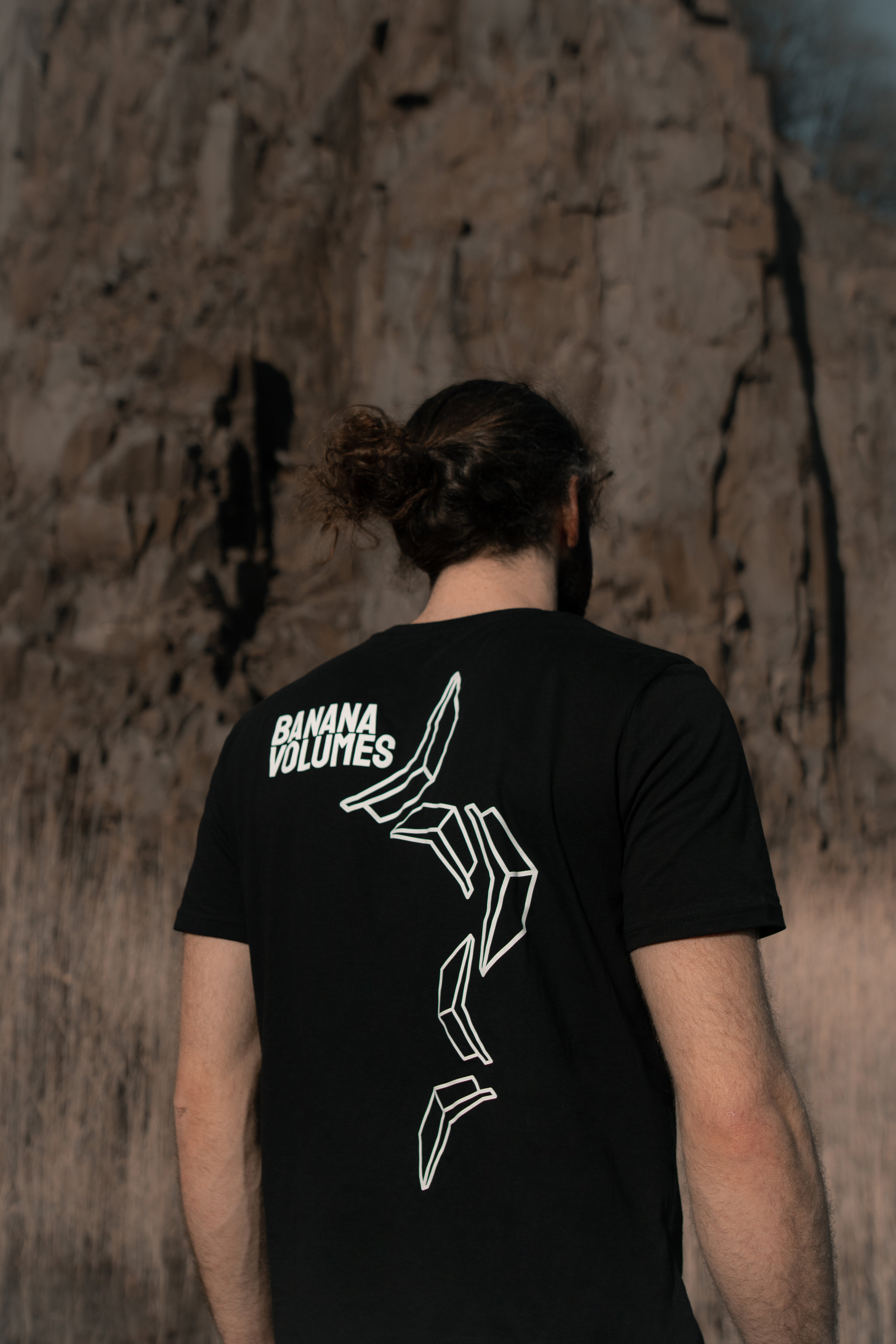Ein Mann in einem schwarzen Banana Volumes T-Shirt steht mit dem Rücken zur Kamera vor einer Felswand.