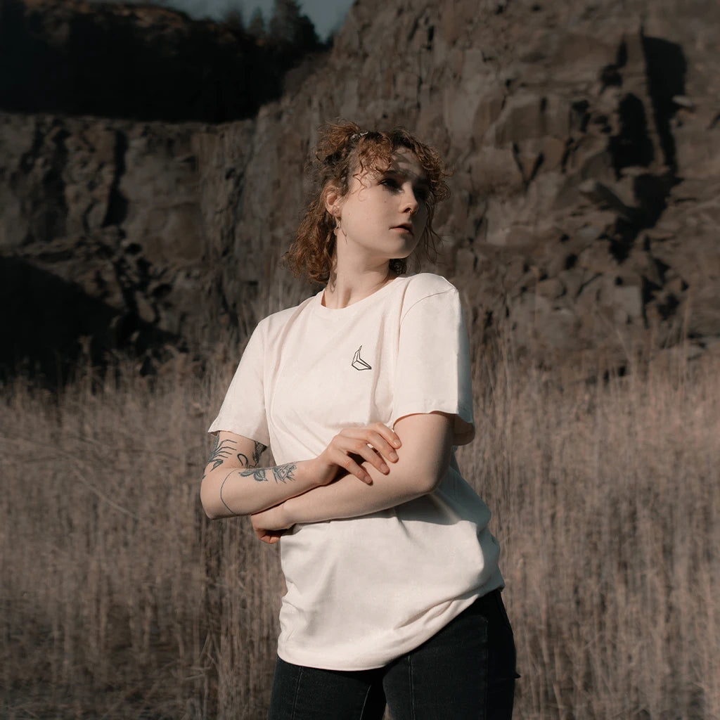 Eine Frau steht vor einer Felswand in einem Feld, sie trägt ein weißes Banana Volumes T-shirt.