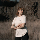 Eine Frau steht vor einer Felswand in einem Feld, sie trägt ein weißes Banana Volumes T-shirt.