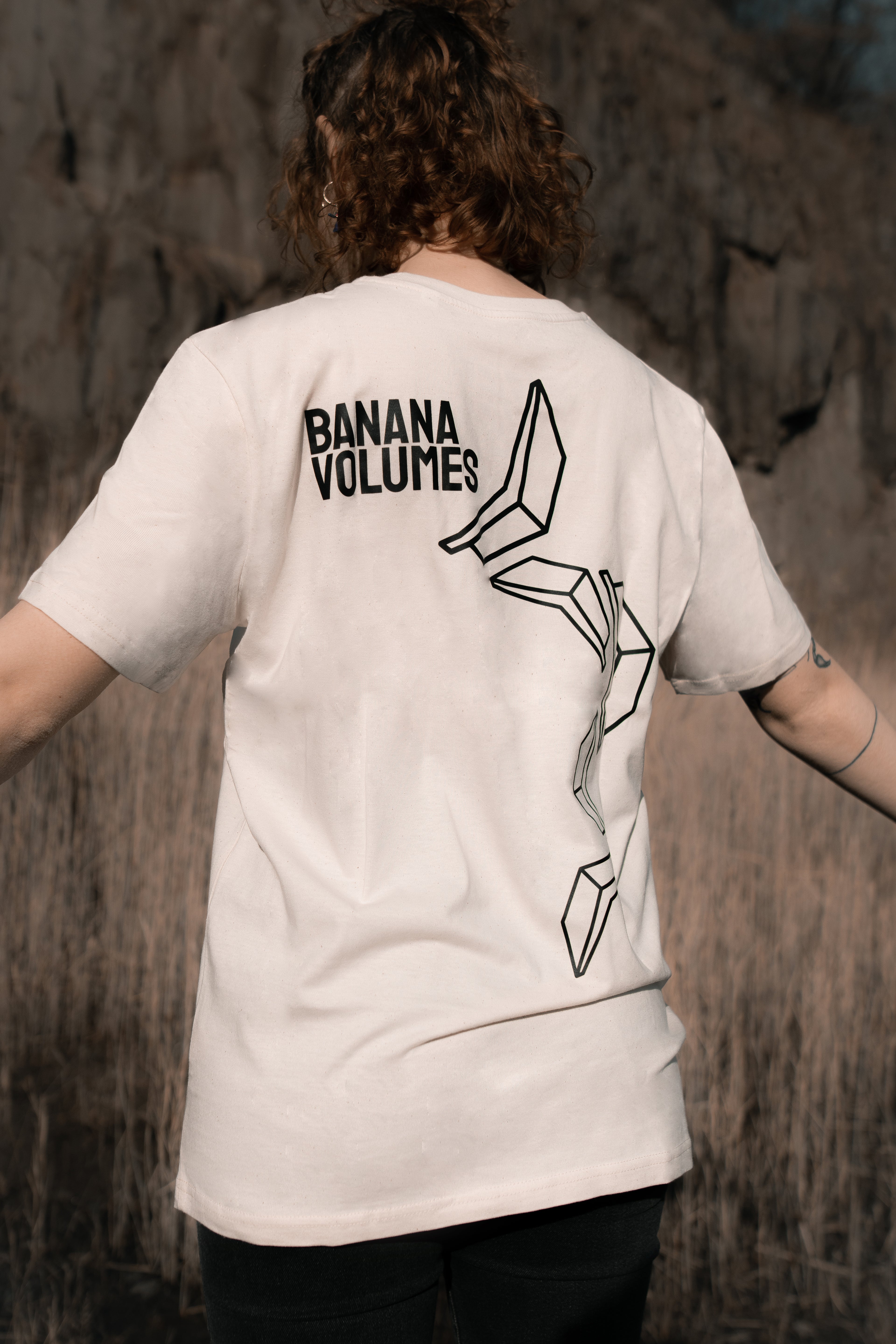 Eine Frau steht mit ausgebreiteten Armen und dem Rücken zur Kamera in einem Feld, sie trägt ein Banana Volumes T-Shirt.