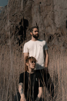 Vor einer Felswand steht ein Mann in einem weißen Banana Volumes T-Shirt  in einem Feld hinter einer sitzenden Frau in einem schwarzen Banana-Volumes T-shirt.