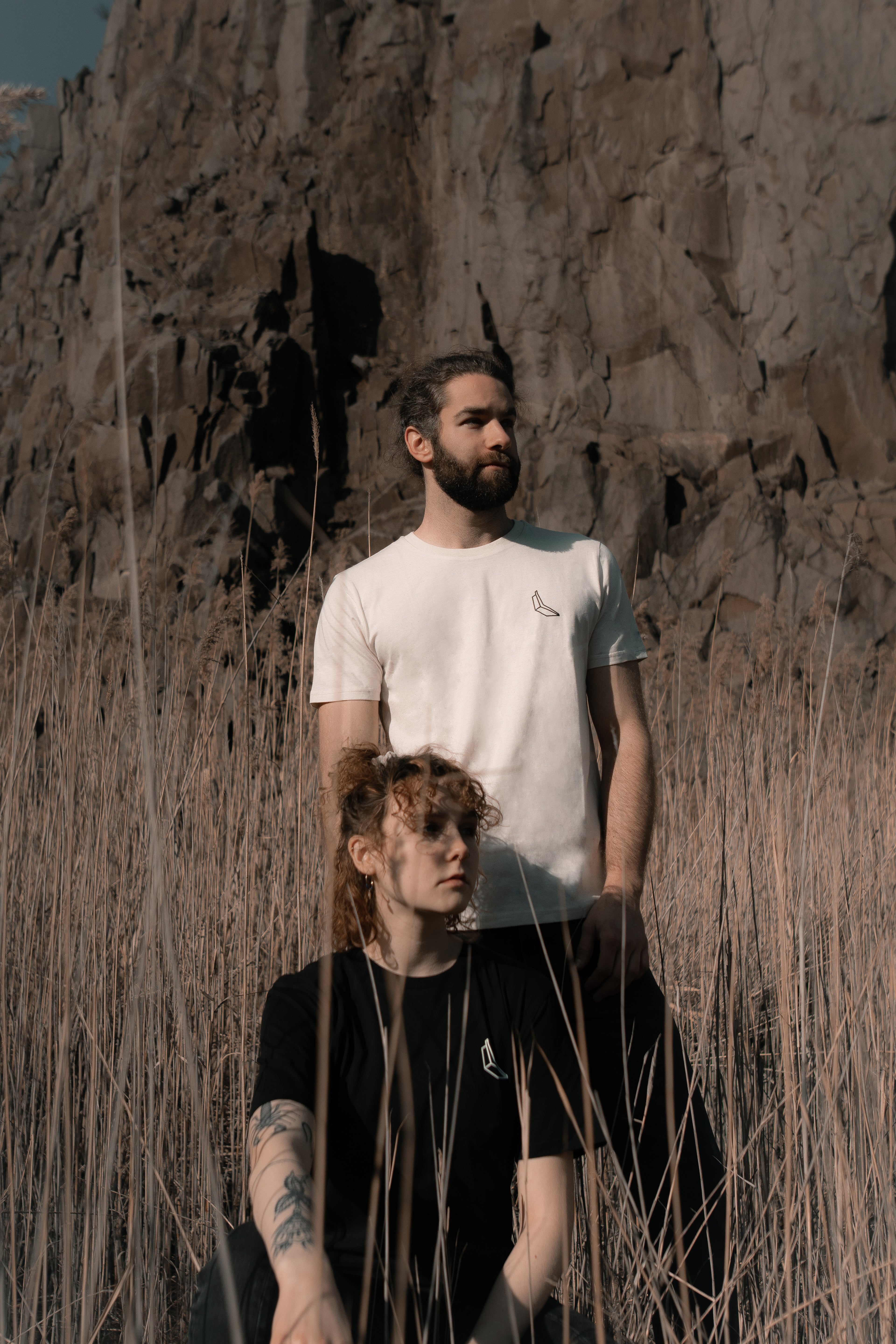 Vor einer Felswand steht ein Mann in einem weißen Banana Volumes T-Shirt  in einem Feld hinter einer sitzenden Frau in einem schwarzen Banana-Volumes T-shirt.