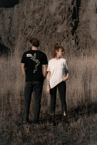 Eine Frau in einem weißen Banana Volumes T-Shirt und ein Mann in einem schwarzen Banana Volumes T-Shirt stehen nebeneinander in einem Feld vor einer Felswand.