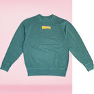 Die Vorderseite eines grünen Sweaters mit Banana Volumes Print in gelb und rosa vor einem rosa-weißen Hintergrund.